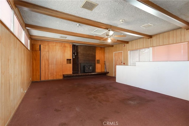 unfurnished living room with wooden walls, beam ceiling, ceiling fan, a textured ceiling, and dark carpet