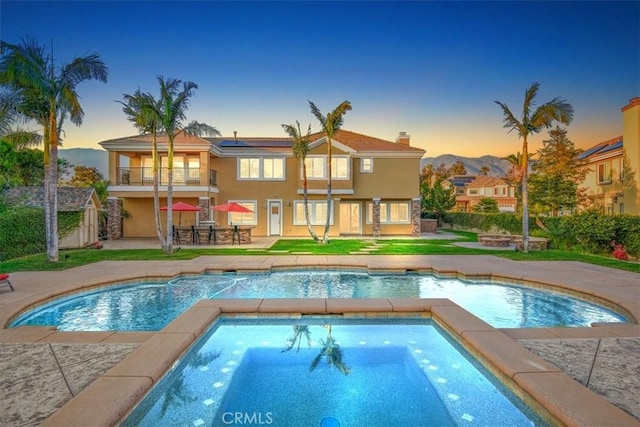 back of property at dusk with a patio, a balcony, solar panels, an in ground hot tub, and stucco siding