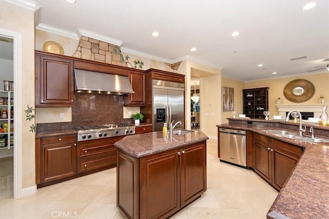 kitchen featuring wall chimney range hood, dark stone counters, an island with sink, stainless steel appliances, and a sink