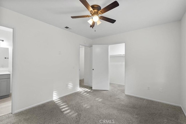 unfurnished bedroom featuring ensuite bath, a walk in closet, light colored carpet, a closet, and ceiling fan