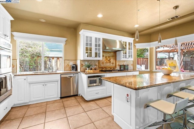 kitchen with wall chimney exhaust hood, pendant lighting, stainless steel appliances, white cabinets, and stone counters