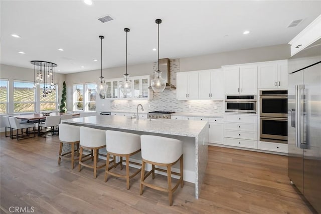 kitchen with appliances with stainless steel finishes, wall chimney range hood, a kitchen island with sink, and white cabinetry