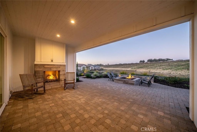 patio terrace at dusk with an outdoor fire pit, an outdoor stone fireplace, and a rural view