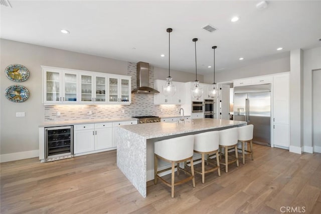 kitchen featuring stainless steel appliances, white cabinets, beverage cooler, a center island with sink, and wall chimney range hood