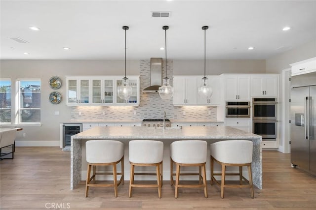 kitchen featuring beverage cooler, stainless steel appliances, an island with sink, white cabinetry, and wall chimney range hood