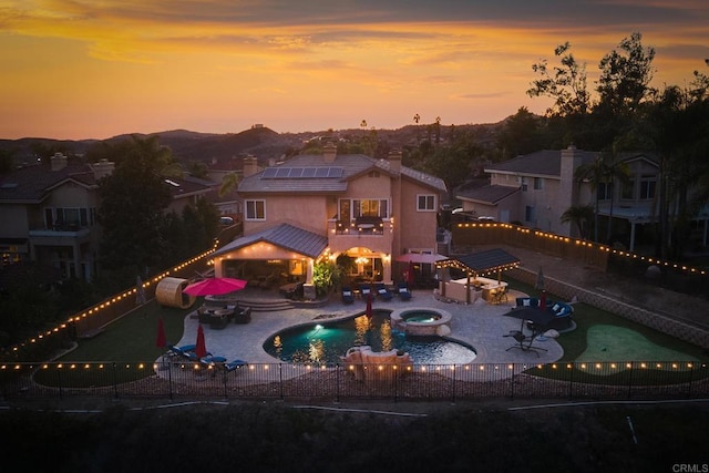 back of property at dusk with roof mounted solar panels, a fenced backyard, a fenced in pool, an in ground hot tub, and a patio area