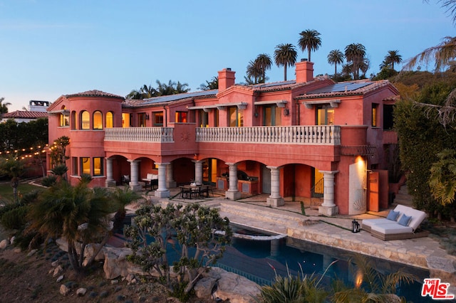 back house at dusk with solar panels, a patio area, and a balcony