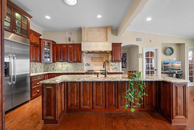 kitchen featuring custom exhaust hood, a center island with sink, stainless steel built in refrigerator, and decorative backsplash