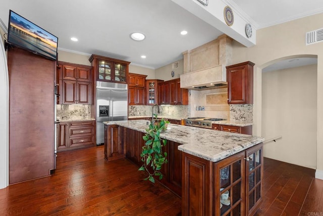 kitchen with stainless steel appliances, light stone counters, tasteful backsplash, an island with sink, and premium range hood