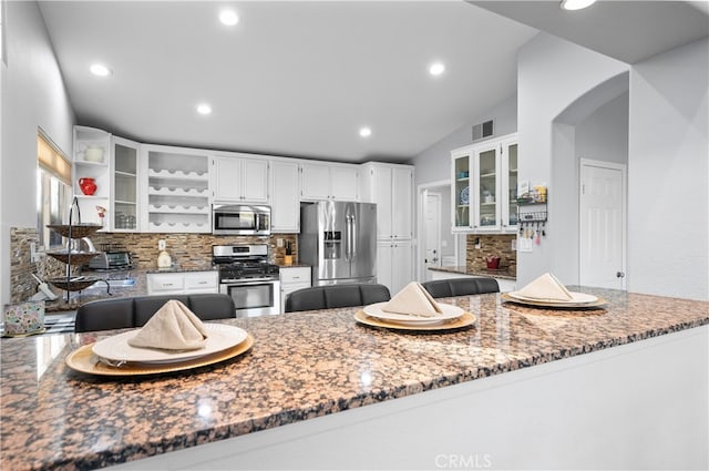 kitchen featuring stainless steel appliances, vaulted ceiling, kitchen peninsula, decorative backsplash, and dark stone counters