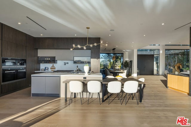 kitchen featuring black double oven, light hardwood / wood-style flooring, hanging light fixtures, a center island with sink, and dark brown cabinetry