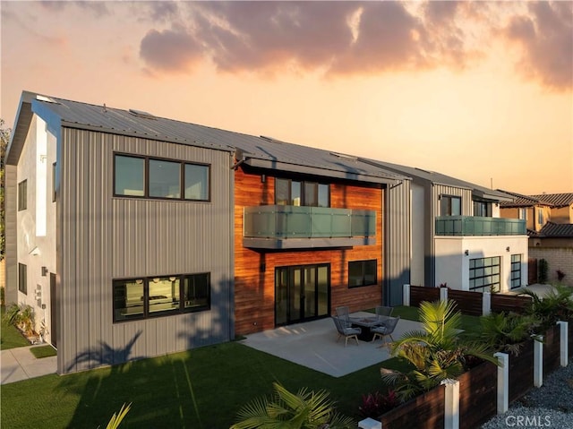 back house at dusk with a balcony, a patio, and a lawn