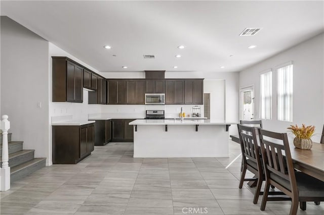 kitchen with a kitchen bar, a kitchen island with sink, dark brown cabinets, and appliances with stainless steel finishes