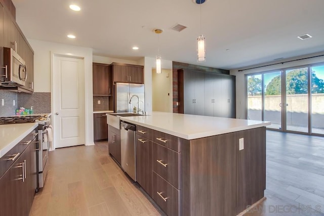 kitchen with stainless steel appliances, light hardwood / wood-style flooring, backsplash, hanging light fixtures, and a kitchen island with sink