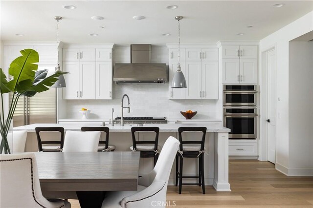 kitchen featuring decorative light fixtures, wall chimney range hood, a center island with sink, and white cabinetry