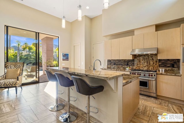 kitchen featuring light brown cabinetry, hanging light fixtures, dark stone countertops, a center island with sink, and high end stove