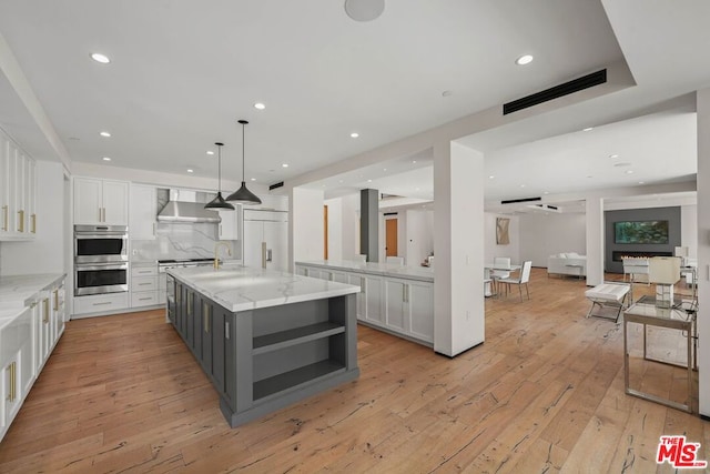 kitchen featuring a large island with sink, wall chimney range hood, stainless steel double oven, light stone countertops, and white cabinets