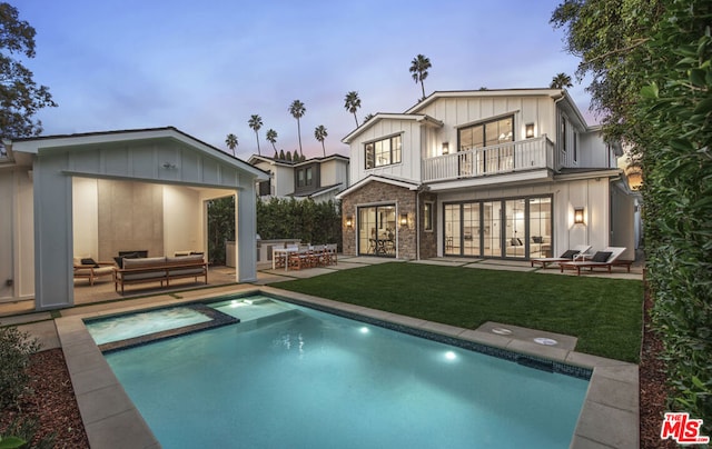 back house at dusk with a yard, a balcony, a pool with hot tub, outdoor lounge area, and a patio area