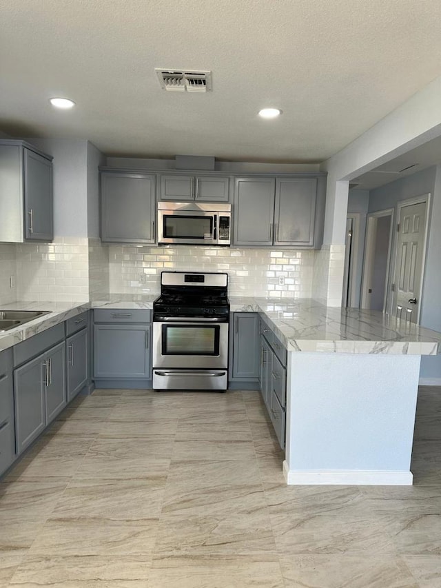 kitchen with gray cabinets, appliances with stainless steel finishes, kitchen peninsula, light stone counters, and decorative backsplash