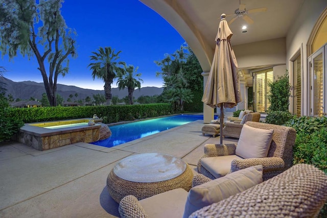 pool at dusk featuring ceiling fan, a patio area, an in ground hot tub, a mountain view, and an outdoor hangout area