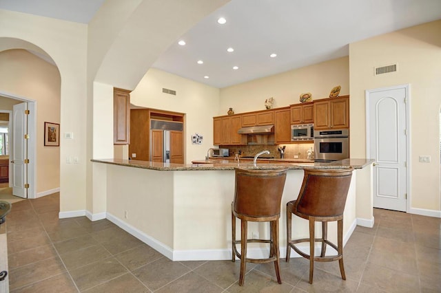 kitchen with kitchen peninsula, built in appliances, light stone counters, a breakfast bar, and sink