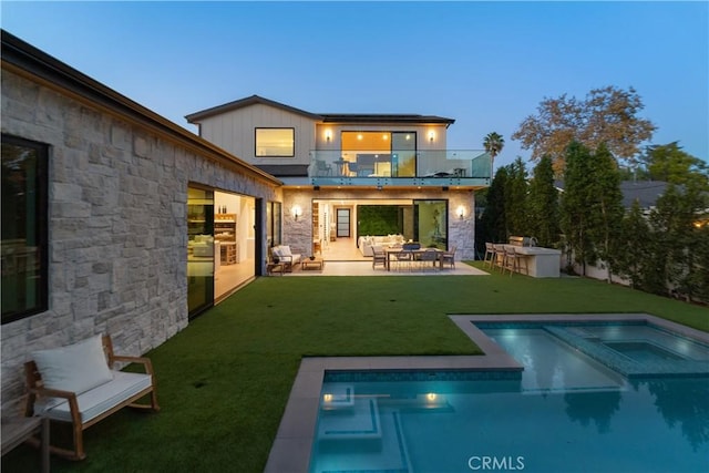 back house at dusk featuring a lawn, an outdoor bar, a balcony, an in ground hot tub, and a patio