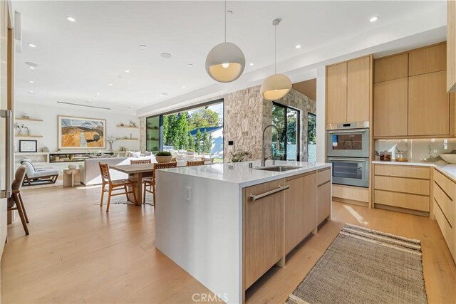 kitchen with a center island with sink, pendant lighting, sink, light brown cabinets, and stainless steel double oven