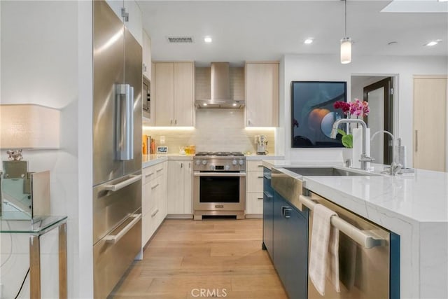 kitchen with tasteful backsplash, high end appliances, wall chimney range hood, decorative light fixtures, and light hardwood / wood-style flooring