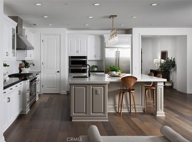 kitchen featuring decorative light fixtures, white cabinetry, high end appliances, wall chimney range hood, and a kitchen island with sink