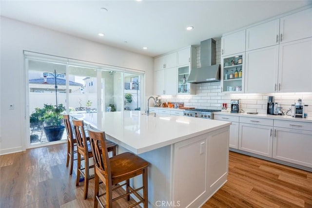 kitchen featuring wall chimney exhaust hood, white cabinetry, a kitchen island with sink, light wood-type flooring, and a breakfast bar area