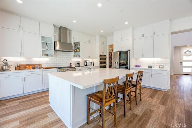 kitchen with white cabinets, a breakfast bar, wall chimney range hood, an island with sink, and stainless steel appliances