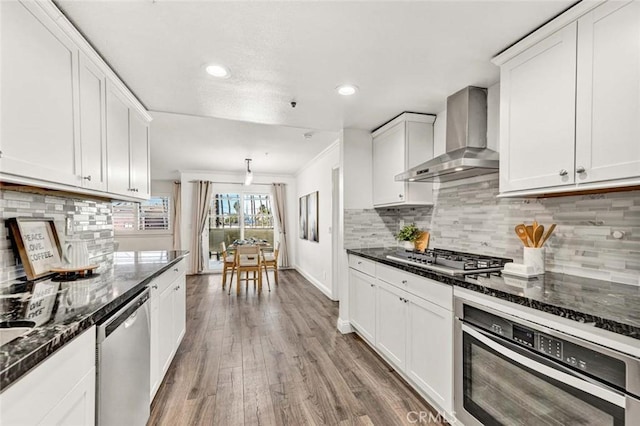 kitchen featuring appliances with stainless steel finishes, wall chimney exhaust hood, wood-type flooring, white cabinetry, and dark stone counters