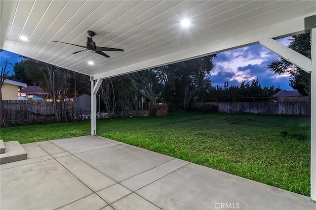 patio terrace at dusk with ceiling fan, a lawn, and a fenced backyard