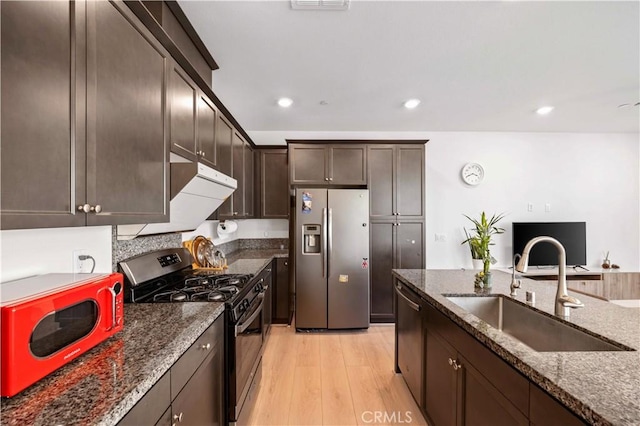 kitchen with appliances with stainless steel finishes, dark stone countertops, dark brown cabinets, light hardwood / wood-style flooring, and sink