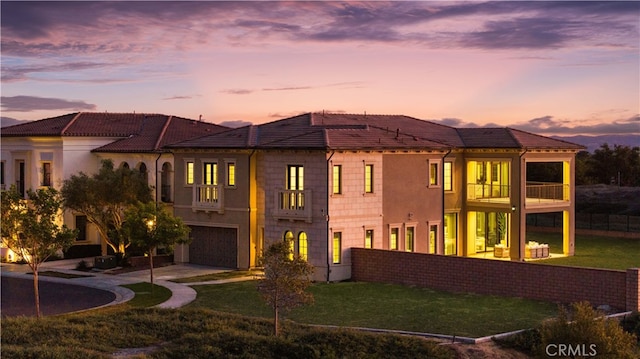 back house at dusk with a garage, a lawn, and a balcony