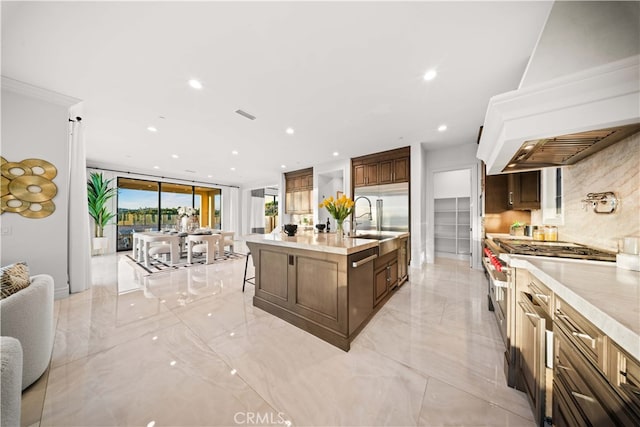 kitchen with backsplash, custom exhaust hood, a kitchen island with sink, stainless steel built in fridge, and a breakfast bar area