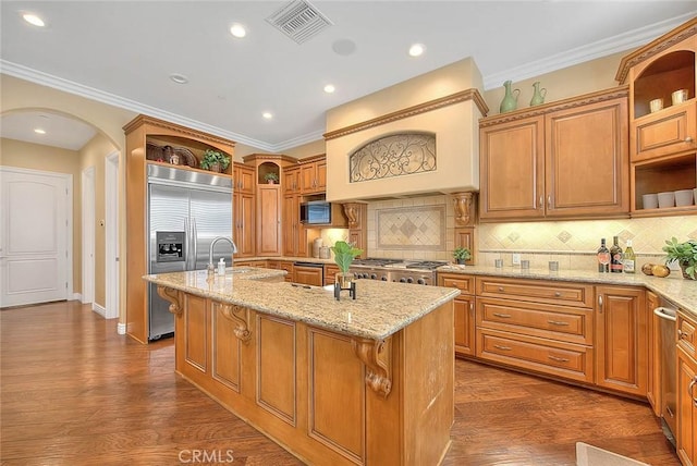 kitchen with ornamental molding, appliances with stainless steel finishes, an island with sink, light stone countertops, and decorative backsplash