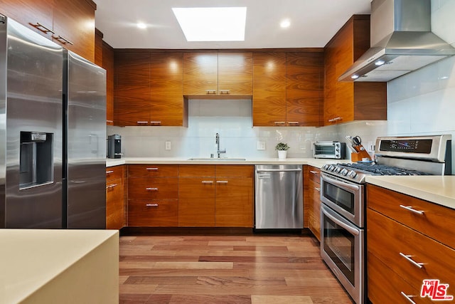 kitchen with appliances with stainless steel finishes, sink, decorative backsplash, wall chimney range hood, and light wood-type flooring