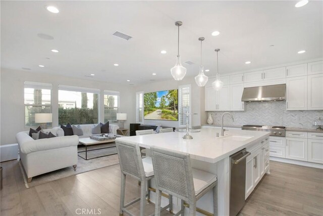 kitchen with decorative light fixtures, wall chimney range hood, sink, an island with sink, and white cabinets