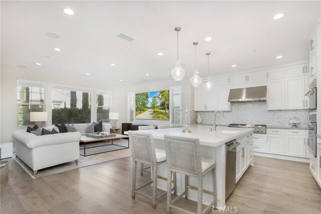 kitchen with wall chimney range hood, pendant lighting, sink, an island with sink, and white cabinets