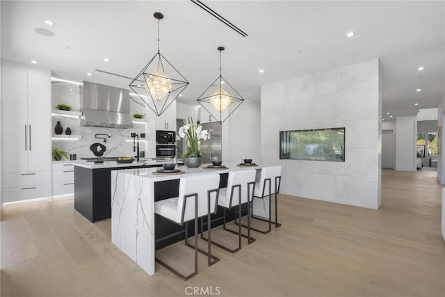 kitchen with hanging light fixtures, white cabinetry, wall chimney range hood, and a center island with sink