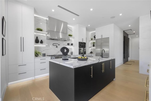 kitchen with white cabinetry, wall chimney exhaust hood, light hardwood / wood-style floors, a kitchen island with sink, and appliances with stainless steel finishes