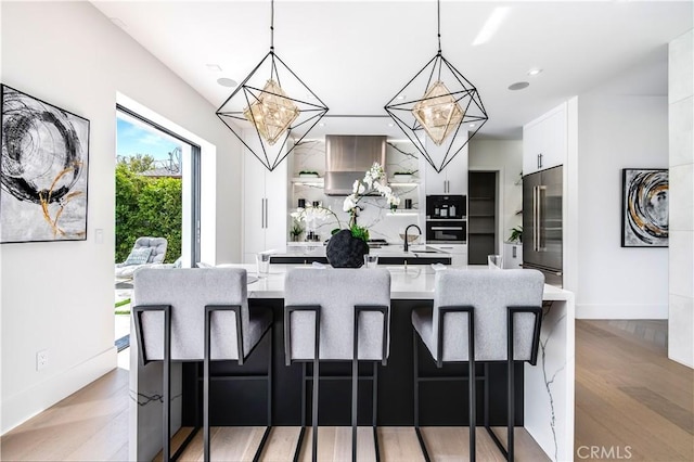 kitchen featuring a kitchen island with sink, stainless steel built in refrigerator, pendant lighting, a kitchen bar, and white cabinetry