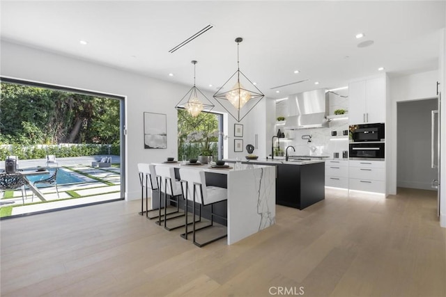 kitchen featuring white cabinets, a kitchen island with sink, wall chimney exhaust hood, and pendant lighting