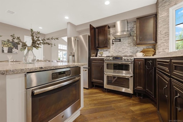 kitchen with backsplash, appliances with stainless steel finishes, wall chimney range hood, and dark brown cabinetry