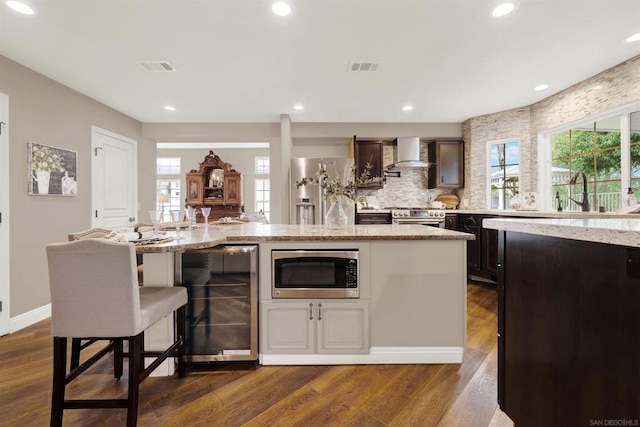 kitchen with a center island with sink, appliances with stainless steel finishes, dark hardwood / wood-style flooring, beverage cooler, and wall chimney exhaust hood