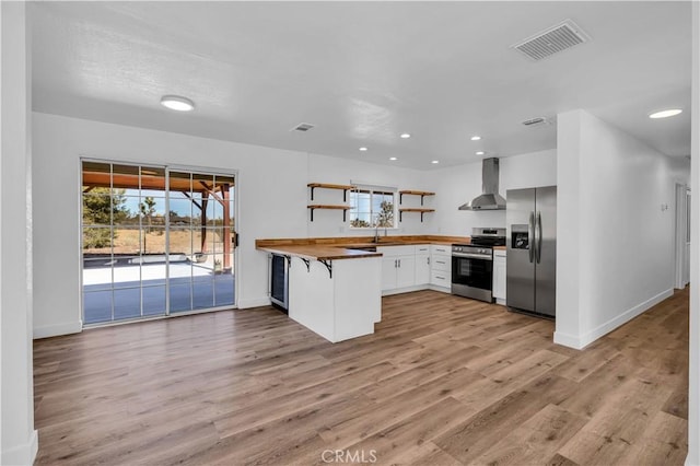 kitchen featuring white cabinets, appliances with stainless steel finishes, wall chimney range hood, butcher block counters, and kitchen peninsula