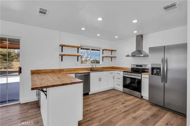 kitchen with appliances with stainless steel finishes, butcher block countertops, sink, white cabinets, and wall chimney exhaust hood