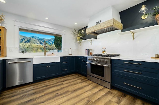 kitchen with appliances with stainless steel finishes, premium range hood, hardwood / wood-style flooring, sink, and blue cabinets