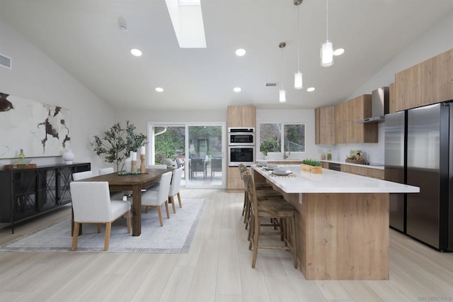 kitchen with appliances with stainless steel finishes, a kitchen island, wall chimney range hood, light hardwood / wood-style floors, and hanging light fixtures